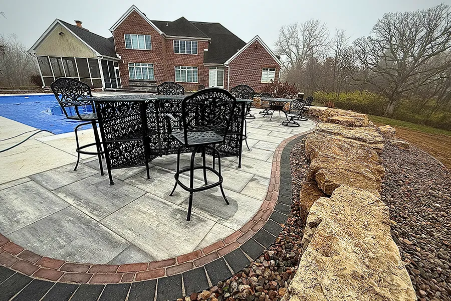 A custom hardscape patio with stone pavers, curved brick edging, outdoor seating, and large boulders bordering the space behind a brick home. - TaylorMade Landworks
