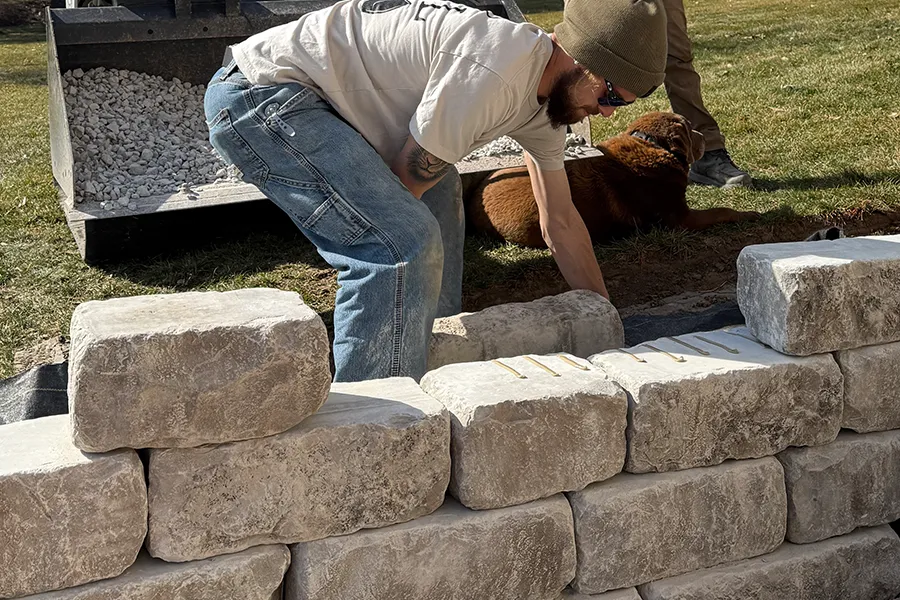 A landscaper placing large stone blocks while building a retaining wall, with gravel and tools visible in the background. - TaylorMade Landworks